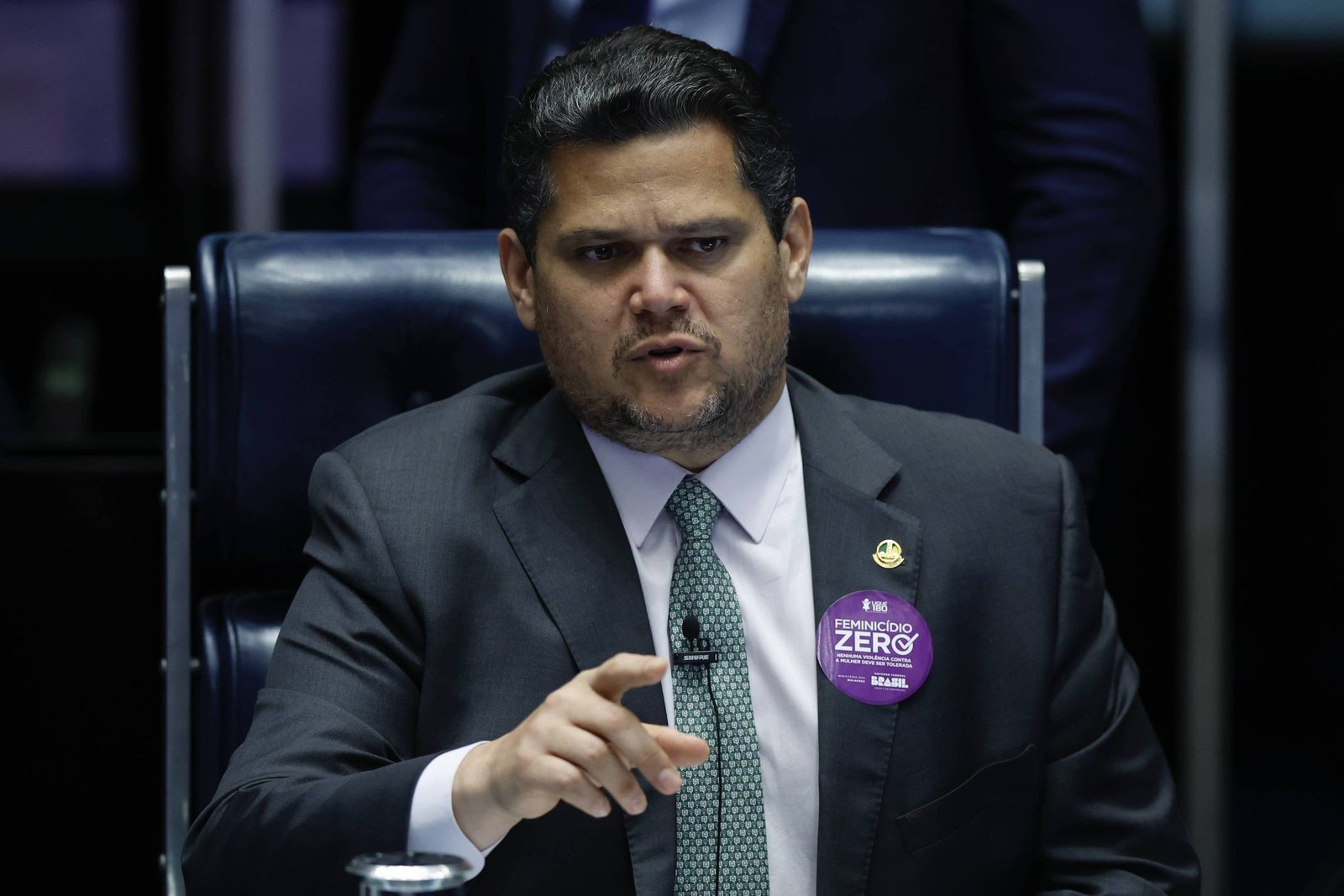 Homem de terno escuro e gravata verde sentado em cadeira azul escura, gesticulando com a mão direita. Ele usa um botão roxo no paletó com texto sobre