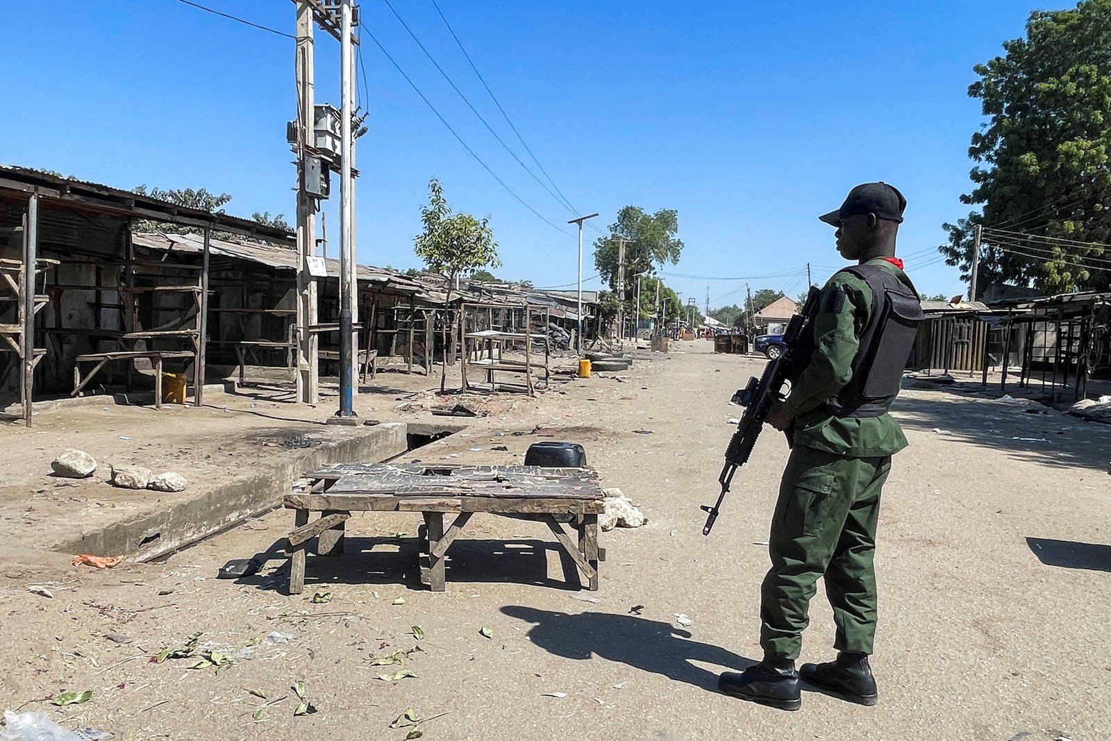 Soldado com uniforme verde e capacete preto segura arma e vigia mercado deserto com barracas fechadas sob céu azul.