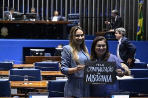 Duas mulheres posam sorrindo no plenário do Senado Federal segurando cartaz preto com texto branco: