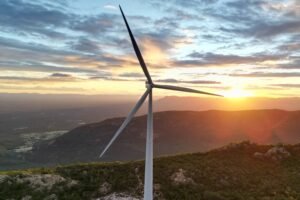 A imagem mostra uma turbina eólica em primeiro plano, com suas lâminas visíveis. Ao fundo, há uma paisagem montanhosa sob um céu colorido, onde o sol está se pondo, criando um efeito de luz dourada. A vegetação é visível nas encostas das montanhas, e o horizonte se estende com uma vista ampla.