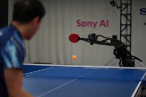 Robô equipado com raquete vermelha em braço mecânico executa movimento para rebater bola laranja em mesa de tênis de mesa. Homem em camiseta azul observa a ação do robô. Fundo com texto parcial