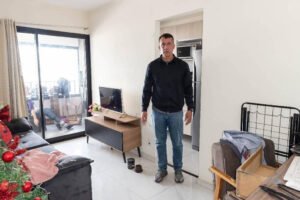 Homem de pé em sala de estar com janela grande ao fundo mostrando vista externa. Ambiente com sofá, mesa de centro, cadeira e árvore de Natal decorada à esquerda.