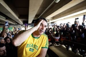 Homem com camisa amarela do Brasil faz gesto de saudação com a mão direita na cabeça, cercado por multidão que fotografa e grava com celulares e câmeras, sob estrutura de concreto.