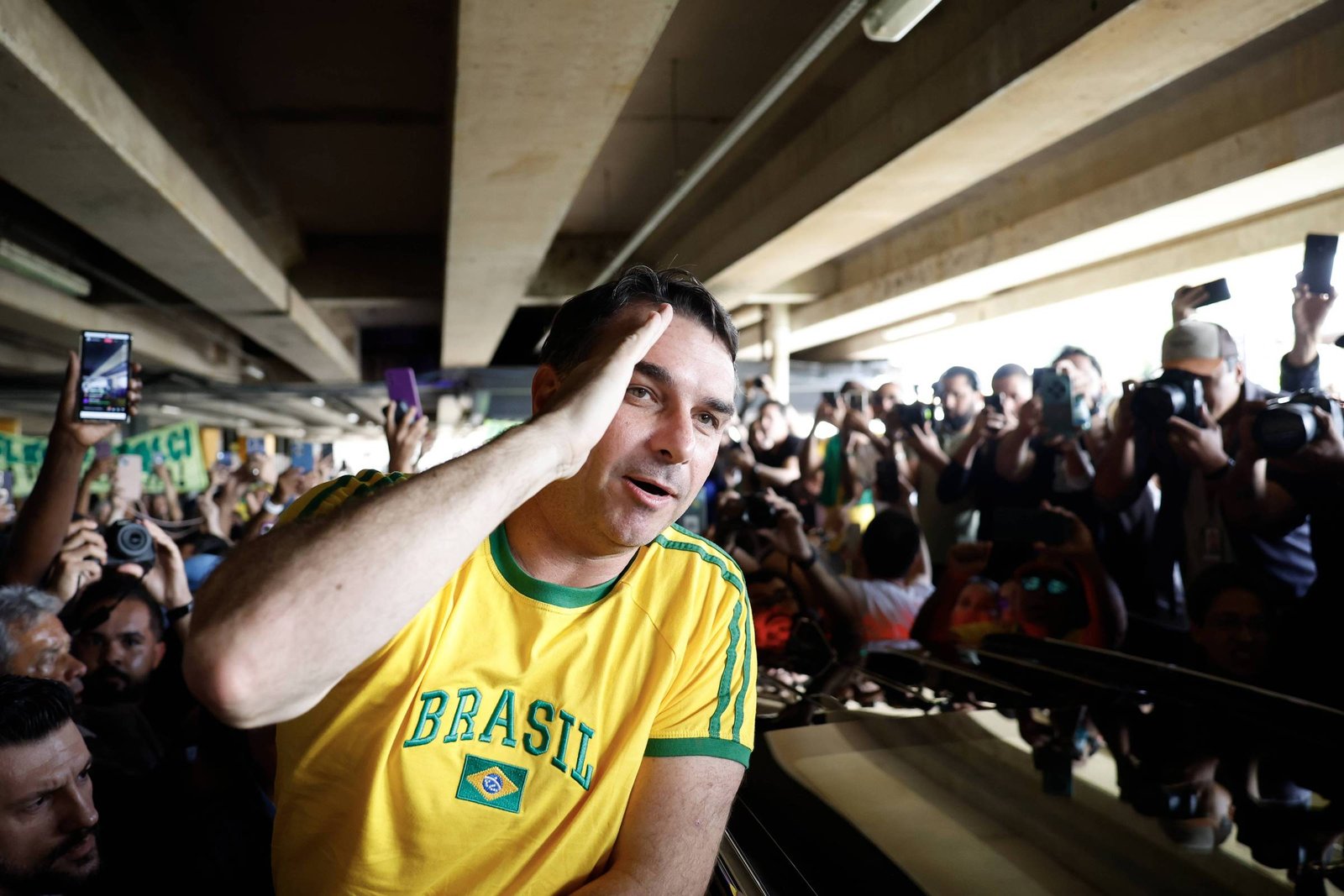 Homem com camisa amarela do Brasil faz gesto de saudação com a mão direita na cabeça, cercado por multidão que fotografa e grava com celulares e câmeras, sob estrutura de concreto.