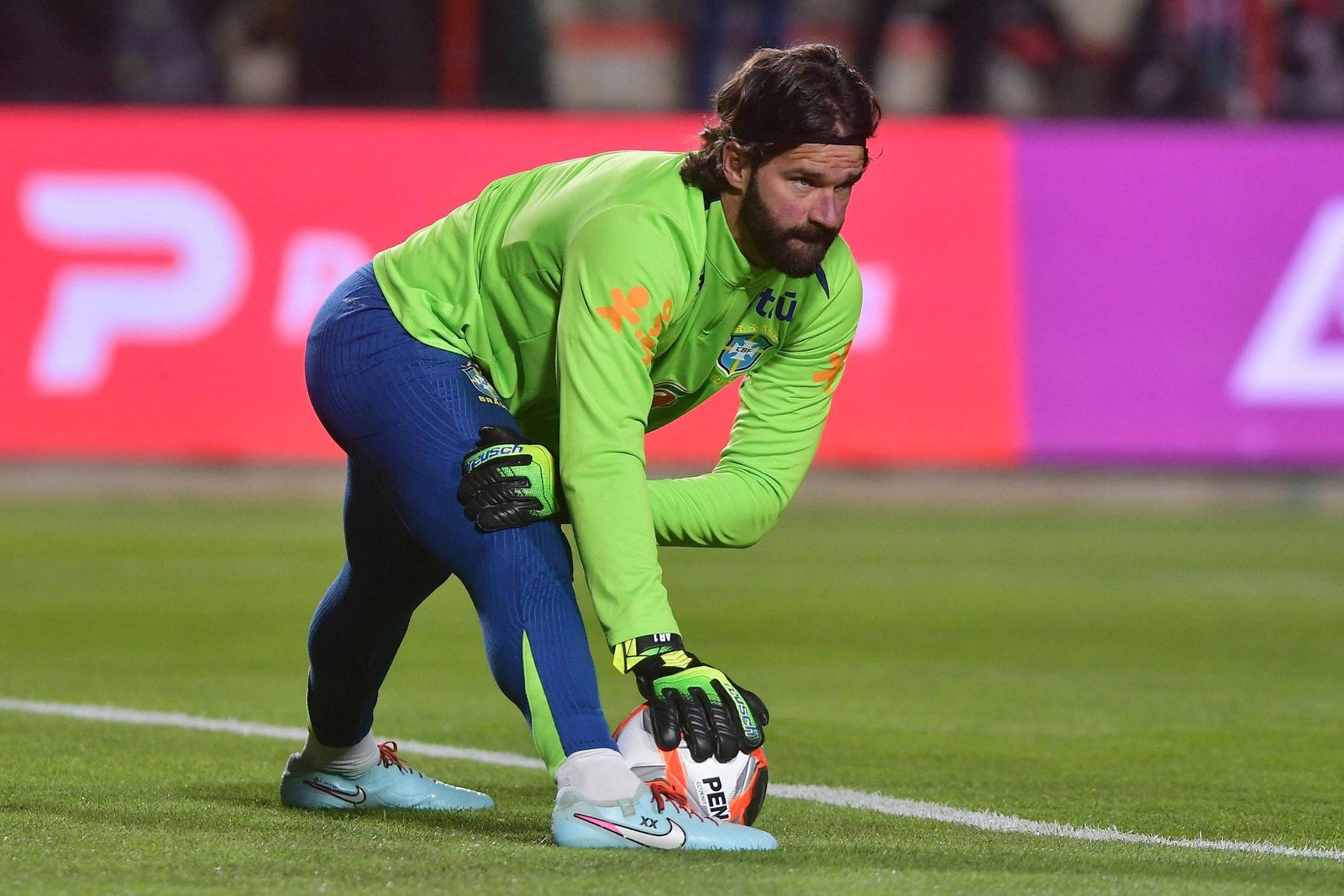 Goleiro com uniforme verde e calça azul agacha-se no campo segurando uma bola de futebol com as duas mãos, focado na ação. Fundo desfocado com publicidade colorida.
