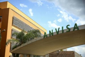 Fachada do prédio da Anvisa em dia claro com céu parcialmente nublado. Letreiro verde com as letras