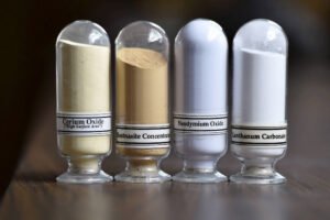 Quatro frascos transparentes alinhados contendo pós de diferentes cores e texturas. Da esquerda para a direita: pó amarelo claro rotulado como Cerium Oxide, pó marrom claro rotulado como Bastnasite Concentrate, pó branco rotulado como Neodymium Oxide e pó branco rotulado como Lanthanum Carbonate. Fundo desfocado e superfície de madeira escura.