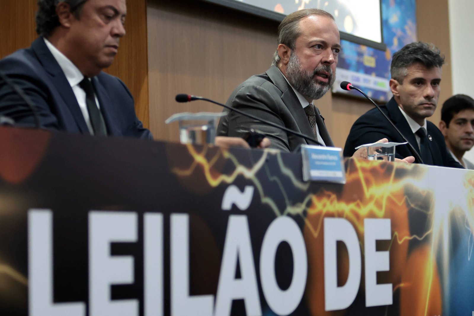 Três homens de terno sentados em mesa com microfones durante coletiva. Banner na frente exibe texto parcial