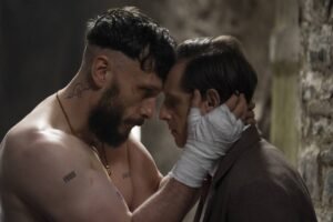 Homem sem camisa com tatuagens e mãos enfaixadas segura o rosto de outro homem vestido com casaco, ambos encostam as testas em um ambiente com parede de pedra ao fundo.