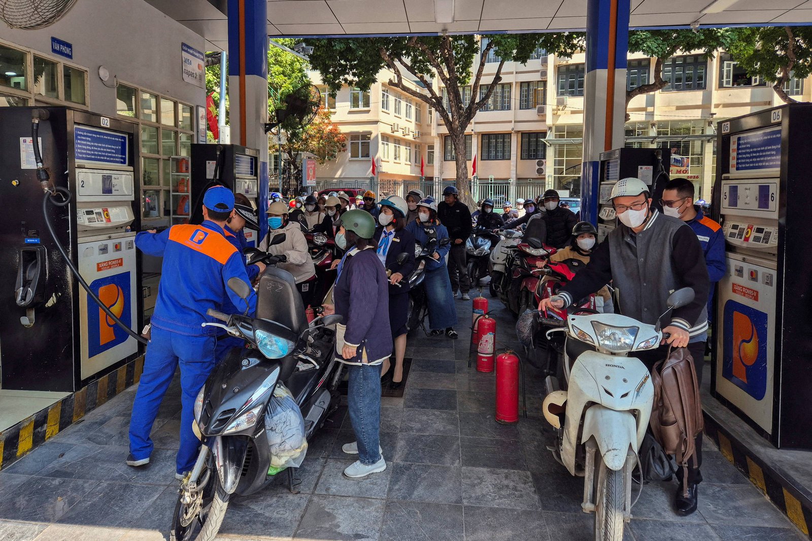 Diversos motociclistas usando capacetes e máscaras formam fila em posto de gasolina durante o dia. Funcionário com uniforme azul e laranja abastece uma moto na bomba à esquerda. Edifícios e árvores aparecem ao fundo.