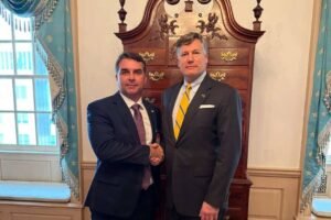 Dois homens de terno escuro apertam as mãos em sala com decoração clássica. Ao fundo, móvel antigo de madeira e duas janelas com cortinas azuis.