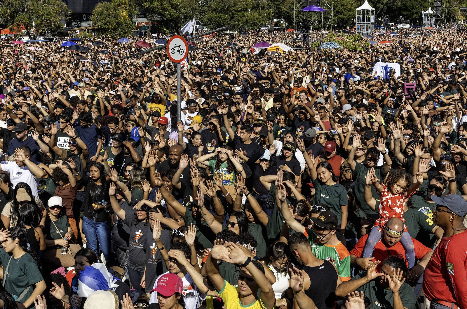 A imagem mostra uma grande multidão reunida em um evento ao ar livre. As pessoas estão levantando as mãos, algumas segurando cartazes. O ambiente é ensolarado, com árvores e tendas ao fundo. Há uma placa de trânsito visível, indicando a proibição de bicicletas. A diversidade de roupas e expressões faciais sugere um evento de grande mobilização.