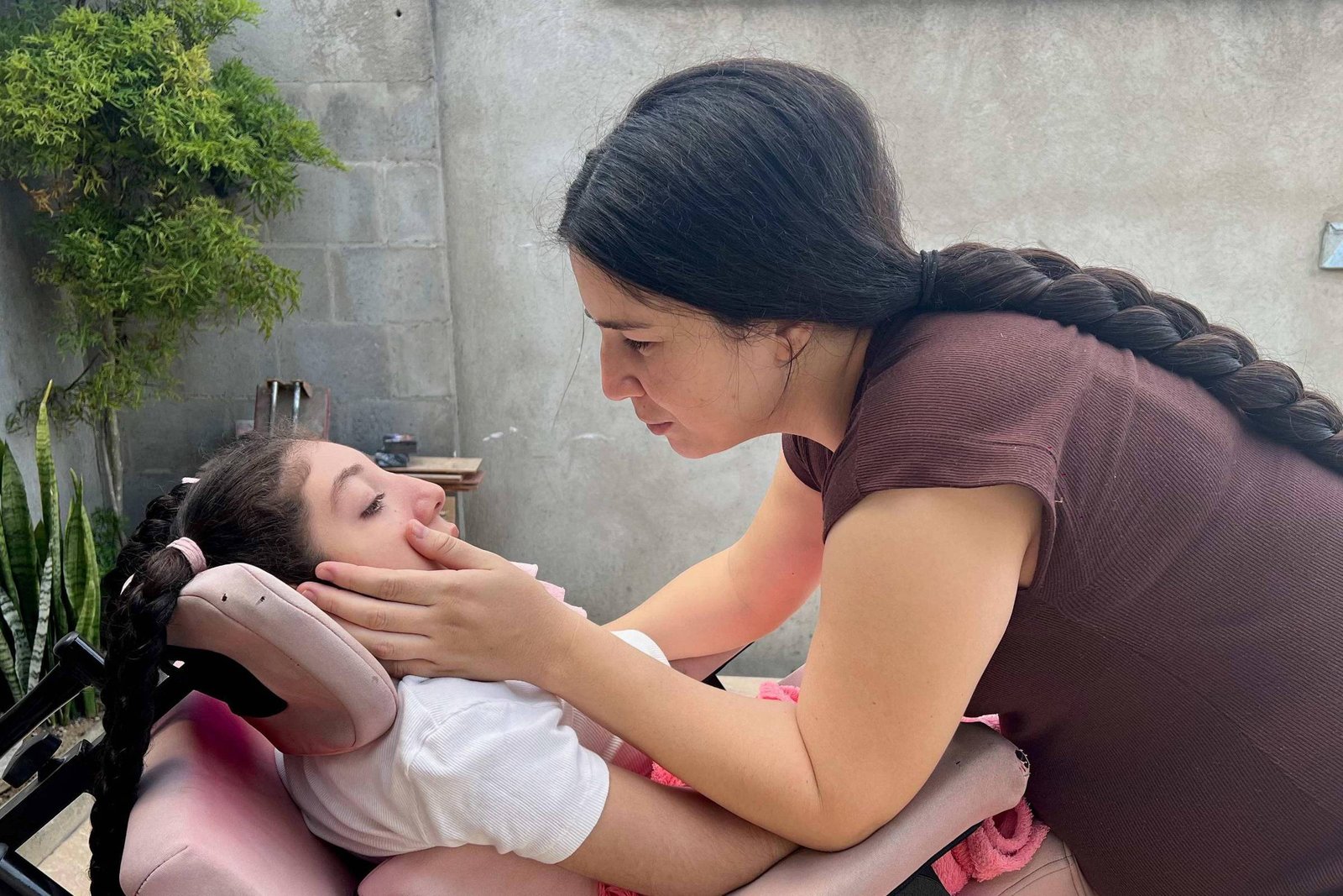 Mulher com cabelo longo e trança segura o rosto de criança reclinada em cadeira de massagem em ambiente externo com plantas e parede cinza.