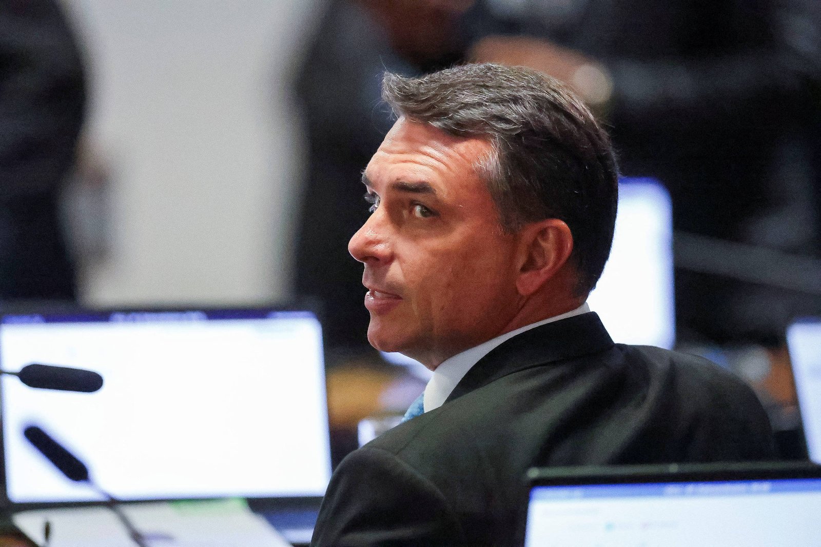 Flávio Bolsonaro, homem de meia-idade com cabelo curto e grisalho veste terno escuro e camisa branca, sentado em frente a telas de computador em ambiente interno, possivelmente escritório ou reunião.