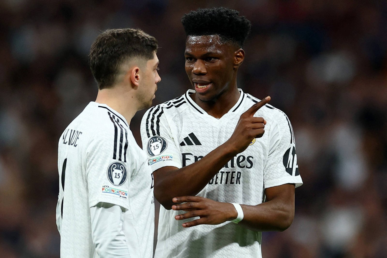 Dois jogadores do Real Madrid em uniforme branco conversam no campo durante jogo. Um deles aponta com o dedo enquanto o outro escuta atentamente. Público desfocado ao fundo.