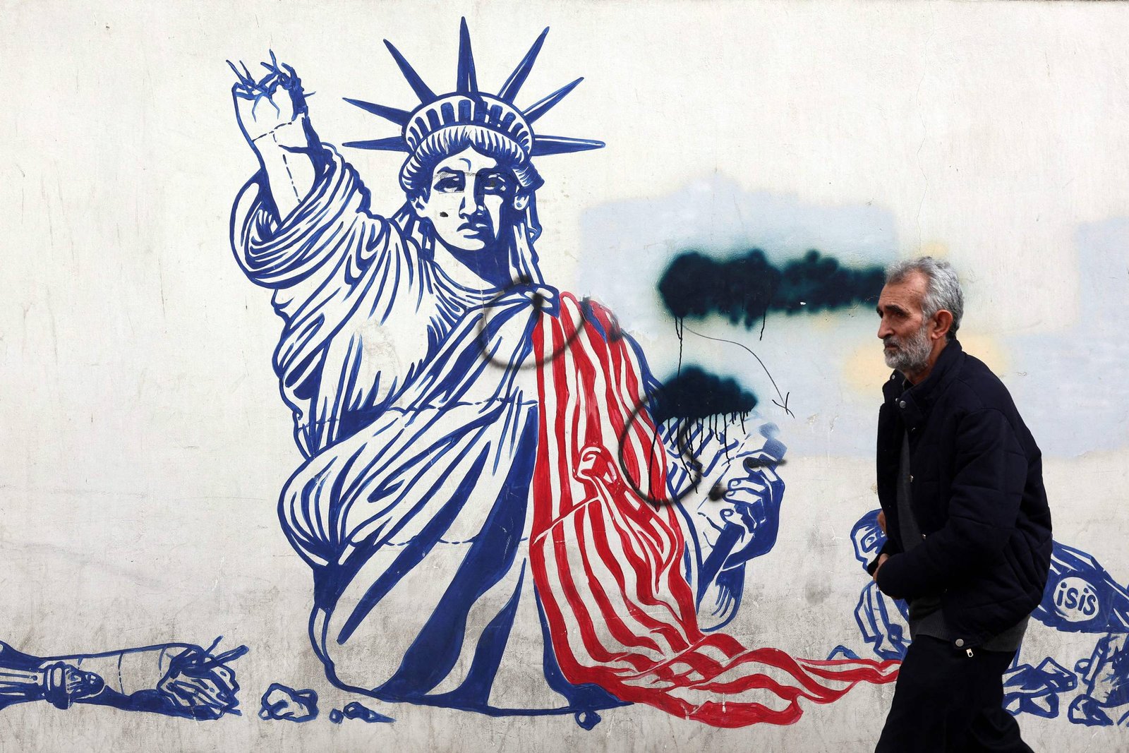 Mural na parede mostra a Estátua da Liberdade parcialmente coberta por uma bandeira dos EUA estilizada em vermelho e branco, com tinta preta borrando parte do rosto. Um homem de cabelos grisalhos e barba caminha à direita, vestindo casaco escuro.