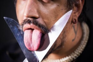 Homem com bigode e barba curta mostra a língua enquanto segura duas facas cruzadas próximas ao rosto, uma delas tocando a língua.