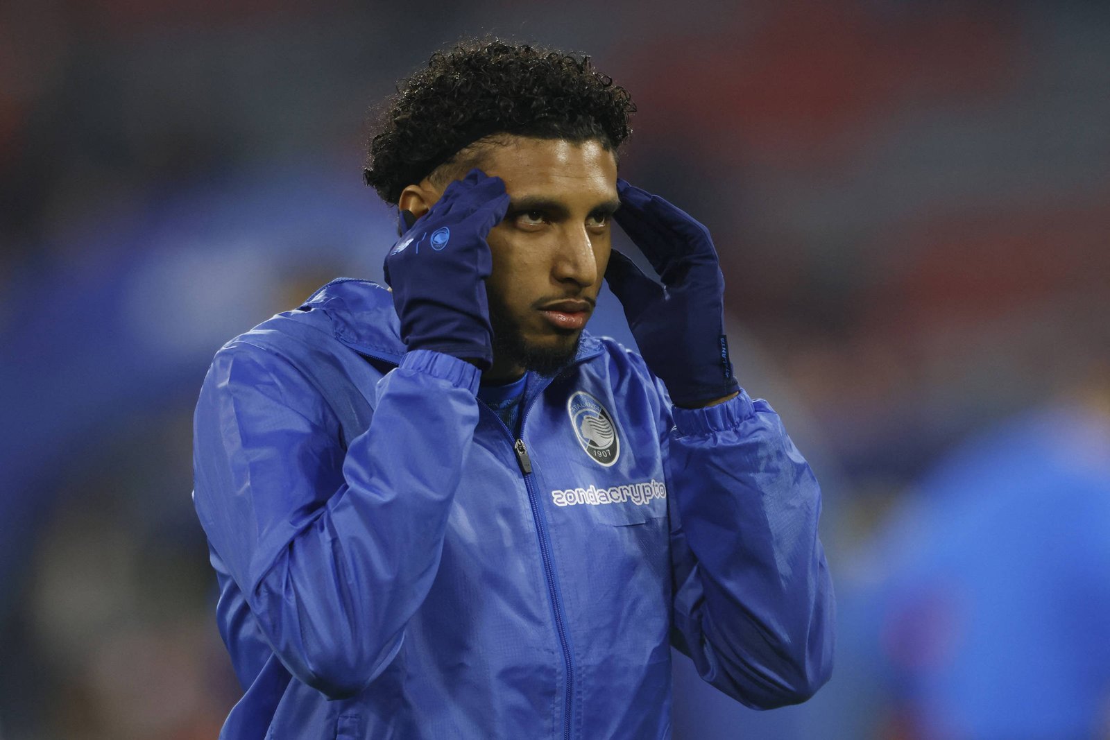 Jogador de futebol com jaqueta azul e luvas ajusta o cabelo antes de partida em estádio com público desfocado ao fundo.