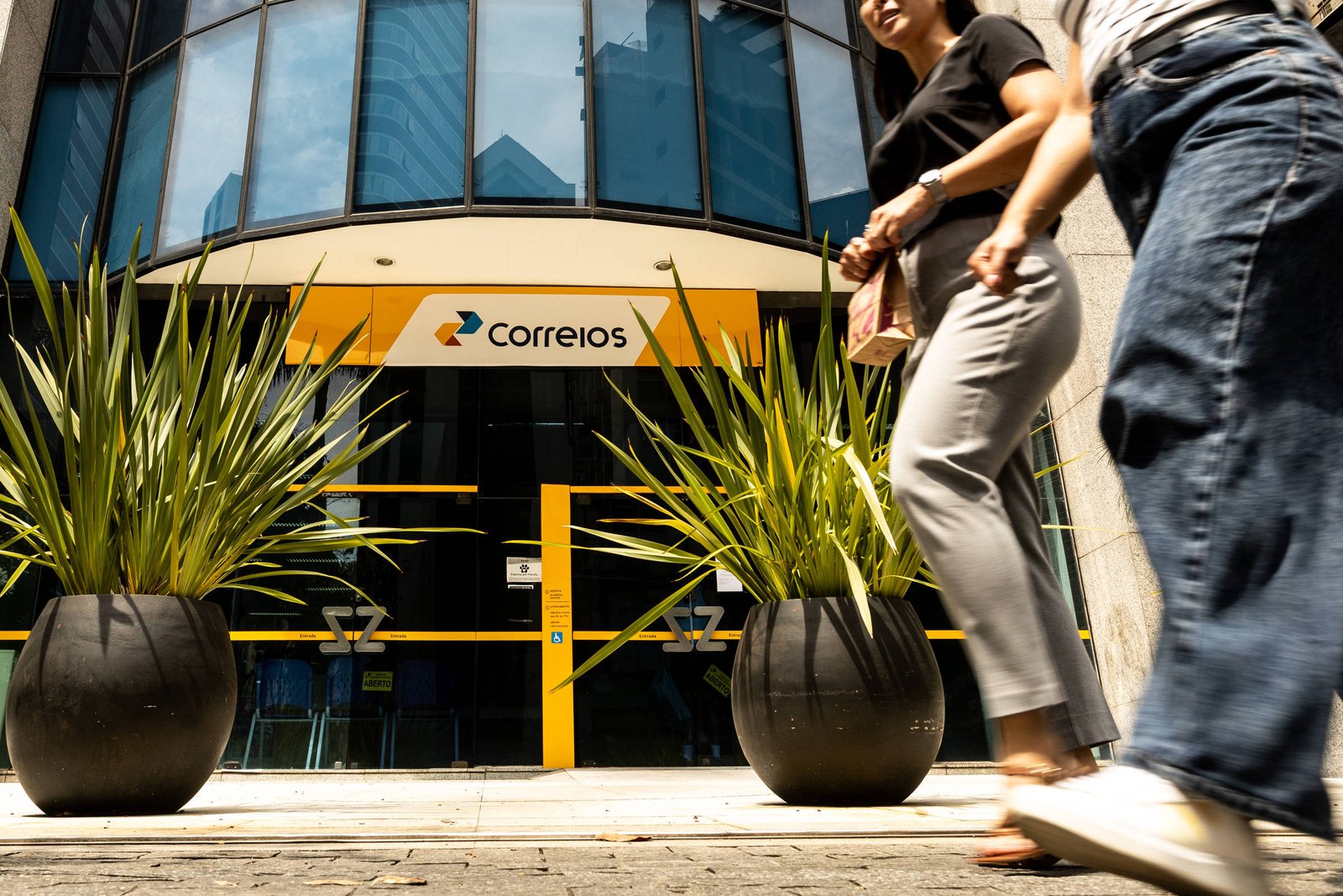 Fachada de agência dos Correios com porta de vidro e detalhes amarelos. Duas plantas grandes em vasos pretos estão posicionadas na calçada. Pessoas caminham em frente ao prédio durante o dia.