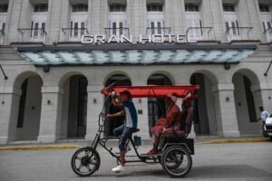 Mototaxi vermelho com motorista e passageiro circula em rua diante de prédio antigo com fachada cinza e janelas altas. Letreiro