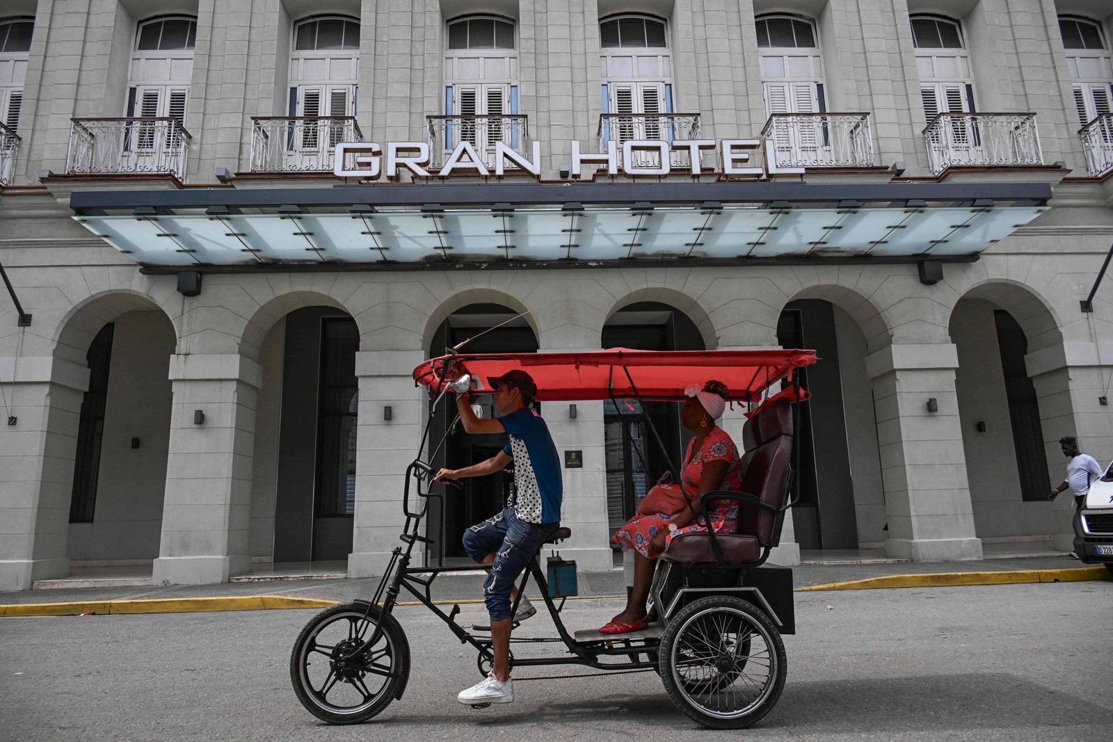 Mototaxi vermelho com motorista e passageiro circula em rua diante de prédio antigo com fachada cinza e janelas altas. Letreiro