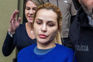 Mulher de cabelo loiro preso e suéter azul com estampa de urso é escoltada por dois policiais civis, um homem à frente e uma mulher atrás, ao sair de um prédio.