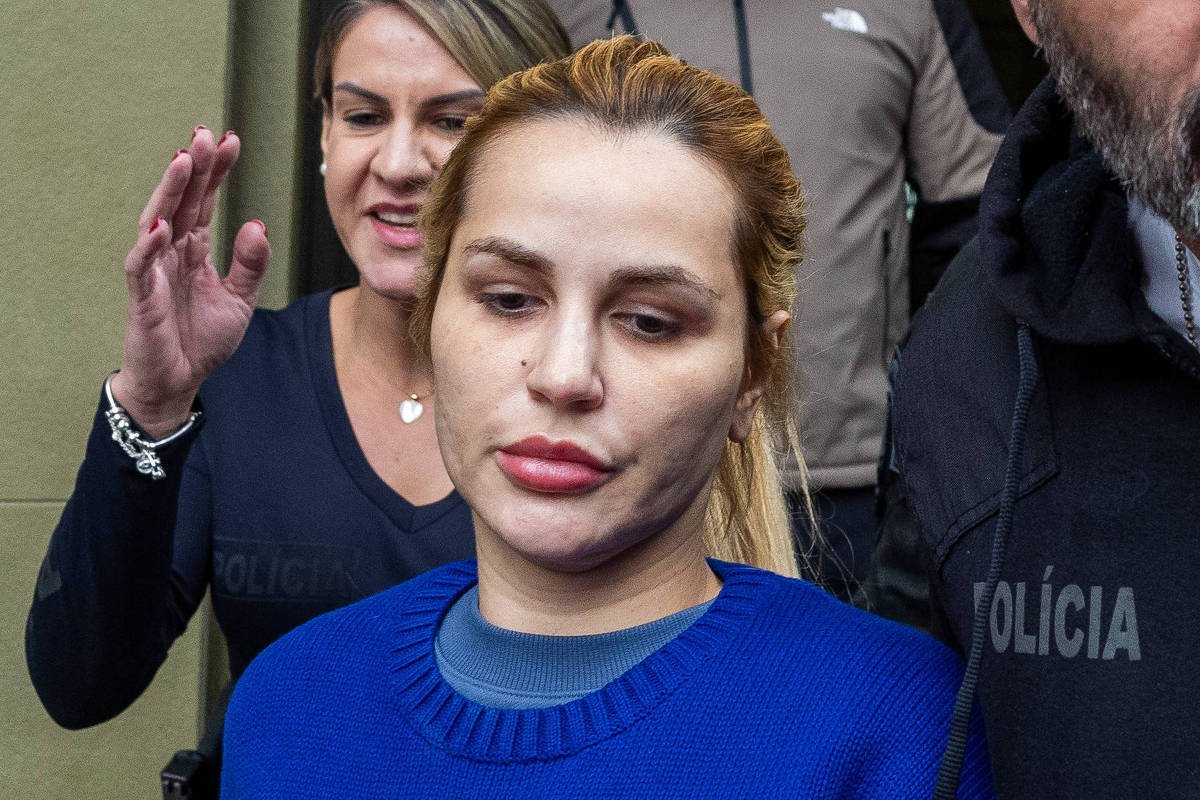 Mulher de cabelo loiro preso e suéter azul com estampa de urso é escoltada por dois policiais civis, um homem à frente e uma mulher atrás, ao sair de um prédio.