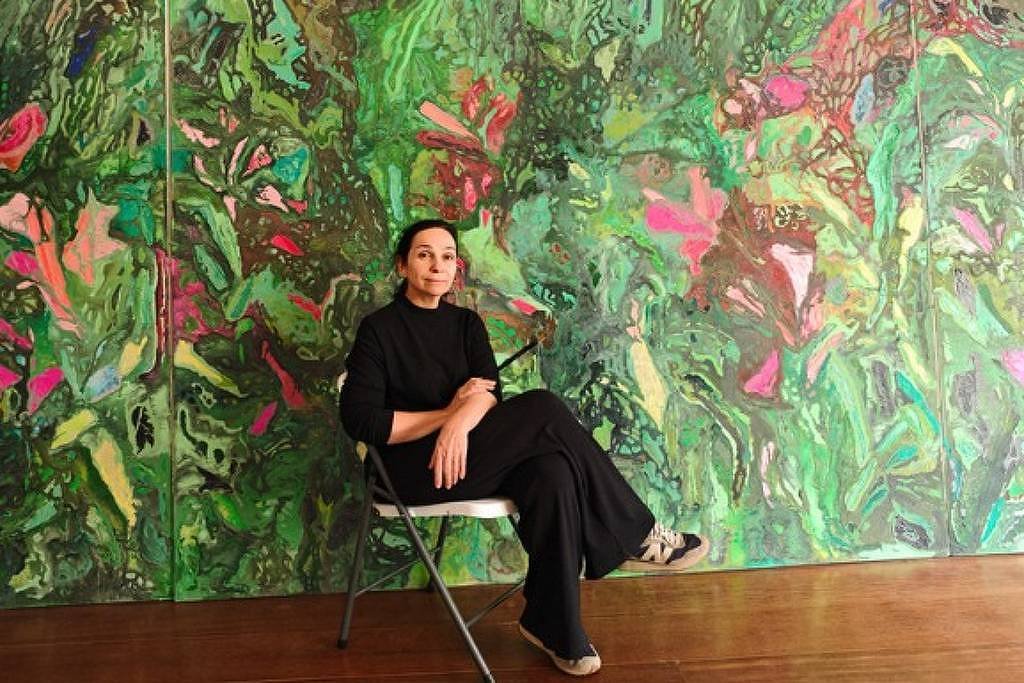 Mulher com roupa preta sentada em cadeira dobrável cinza, com pernas cruzadas, diante de grande pintura abstrata com predominância de verdes, rosas e vermelhos. Piso de madeira clara e parede coberta pela obra vibrante.