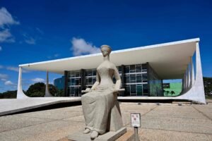 A imagem mostra o Supremo Tribunal Federal (STF) do Brasil, um edifício moderno com uma estrutura de concreto e vidro. Em primeiro plano, há uma escultura de uma figura feminina, representando a Justiça, que está sentada e usa uma venda nos olhos. O céu está claro e azul, com algumas nuvens.