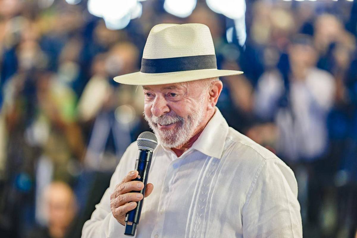 Homem idoso com barba branca usa chapéu branco com faixa preta e camisa social branca enquanto segura microfone. Fundo desfocado mostra várias pessoas e câmeras, sugerindo evento com público.