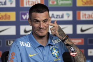 Jogador da seleção brasileira veste camisa azul com emblema da CBF e patrocínios. Ele está sentado em frente a microfone, fazendo gesto com a mão direita perto do olho esquerdo. Fundo com painéis de patrocinadores visíveis.