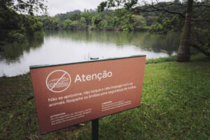 Placa marrom com texto em português e inglês alerta para não se aproximar, tocar ou interagir com animais, em área gramada próxima a lago cercado por árvores e vegetação densa.