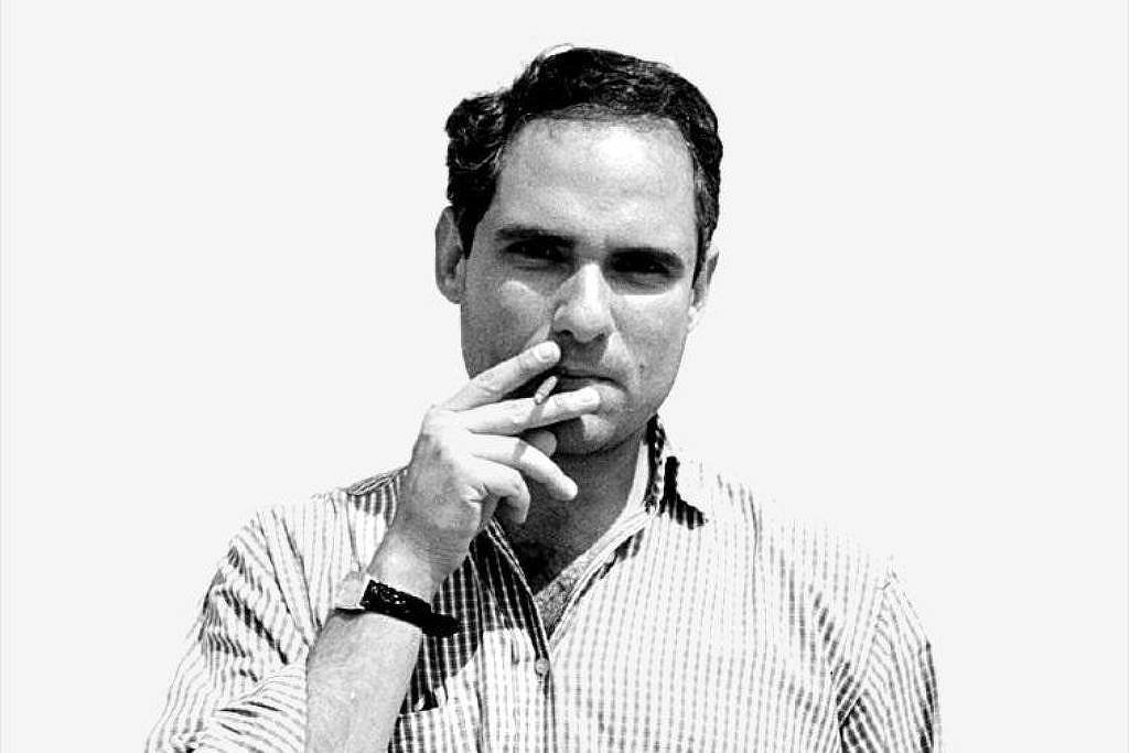 Homem de meia-idade com cabelo curto e escuro, vestindo camisa xadrez de mangas compridas arregaçadas, fuma um cigarro com a mão direita próxima à boca. A mão esquerda está no bolso da calça escura. Fundo branco liso.