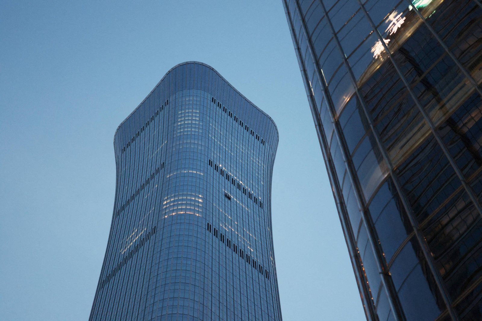 Dois prédios comerciais altos com fachadas de vidro refletem a luz do entardecer. O prédio à esquerda tem formato curvo e janelas iluminadas, enquanto o da direita mostra reflexos do céu.