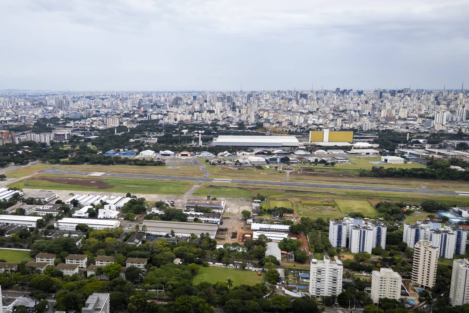 Vista aérea mostra o Campo de Marte com pista de pouso e hangares, cercado por áreas verdes e prédios residenciais. Ao fundo, a extensa malha urbana de São Paulo sob céu nublado.