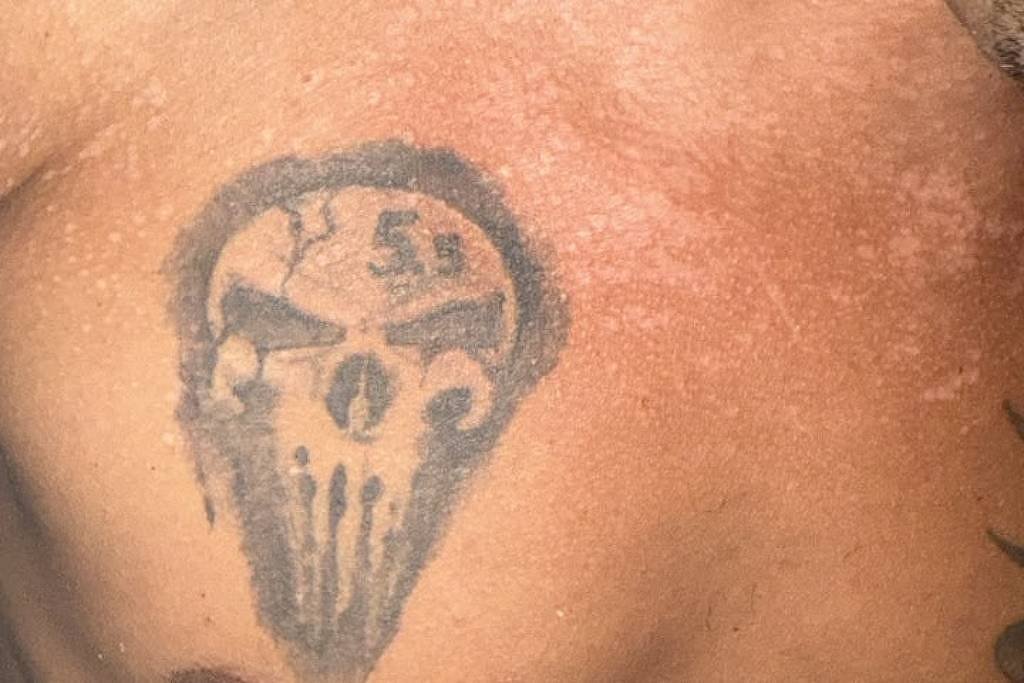 Tatuagem preta de caveira estilizada no lado esquerdo do peito de homem com pele morena. A imagem mostra parte do ombro e do tórax, com iluminação direta.