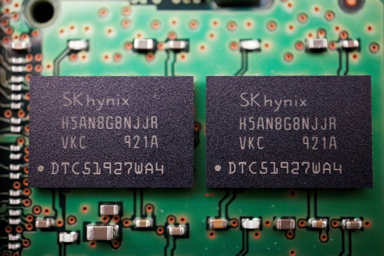 Dois chips pretos SK hynix com inscrições idênticas estão montados lado a lado em uma placa de circuito verde com componentes eletrônicos ao redor.