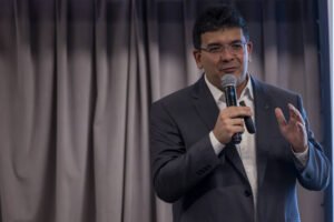 Um homem está em pé, segurando um microfone, enquanto fala. Ele usa um terno escuro e uma camisa branca. O fundo é composto por cortinas cinzas. O homem parece estar explicando algo, gesticulando com a mão livre.