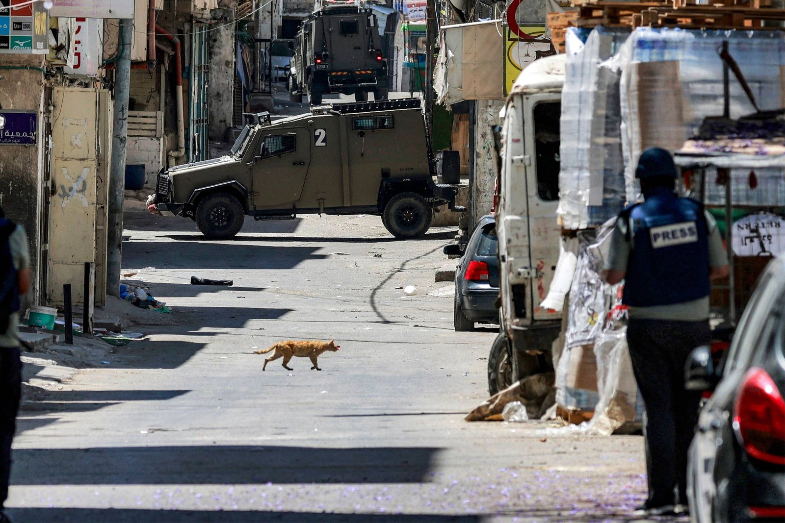 Veículo blindado militar bloqueia rua estreita em área urbana com prédios antigos. Um gato atravessa a rua deserta. À direita, pessoa com colete azul escrito