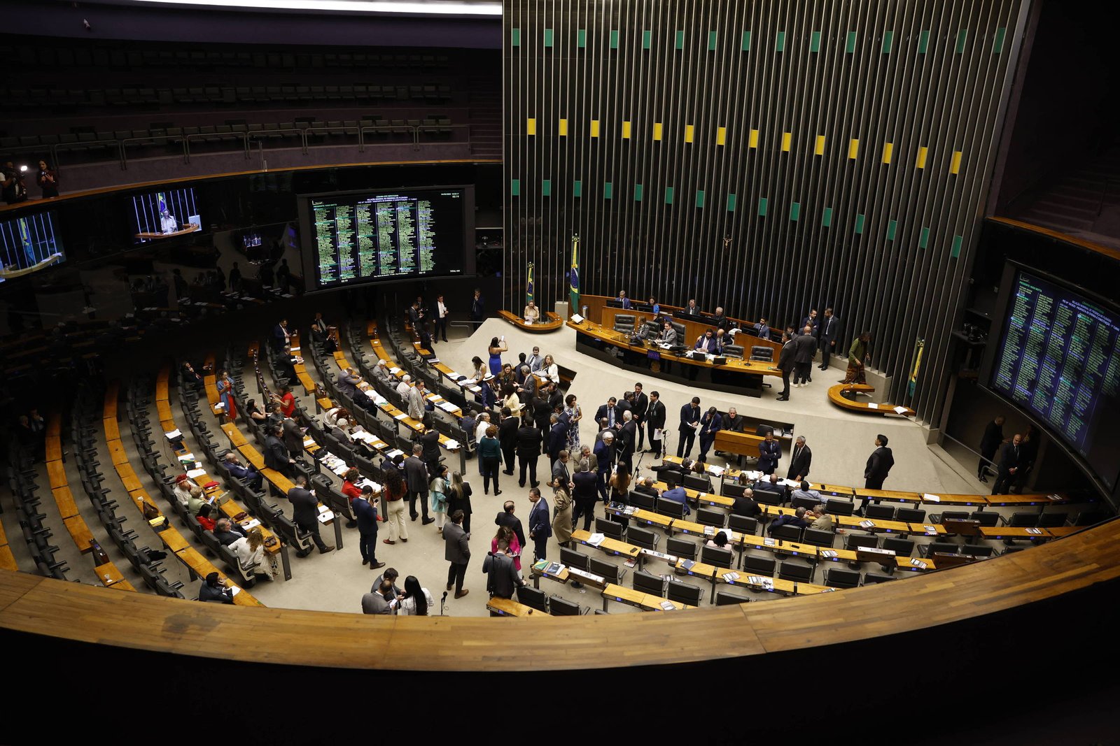 Plenário da Câmara dos Deputados com deputados sentados e em pé, alguns reunidos no centro. Painéis eletrônicos exibem informações legislativas. Ambiente interno amplo com cadeiras organizadas em semicírculo.