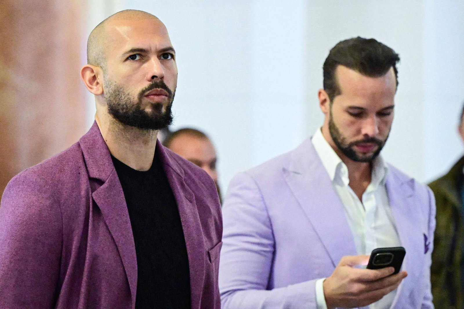 Dois homens estão em ambiente interno. O homem à esquerda é careca, tem barba e veste blazer roxo com camiseta preta. O homem à direita tem cabelo escuro penteado para trás, barba e veste blazer lilás claro com camisa branca, olhando para um celular na mão.