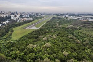 Pista de pouso longa e estreita cercada por densa área verde com árvores. Ao fundo, prédios altos da cidade de São Paulo sob céu nublado.