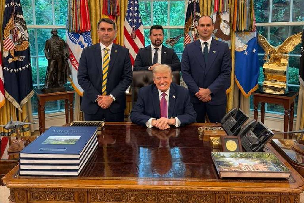 Homem sentado na mesa presidencial do Salão Oval, com três homens em pé atrás dele. Ao fundo, janelas com cortinas douradas, várias bandeiras americanas e estátuas decorativas.