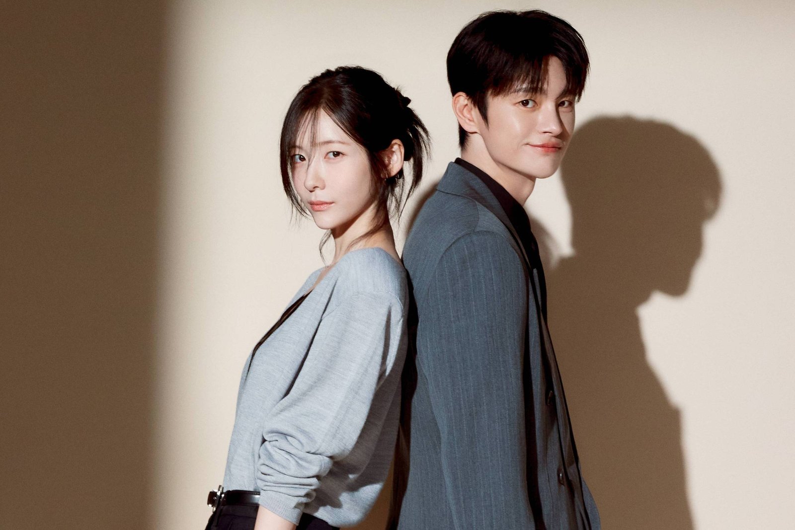 Um homem e uma mulher jovens estão de pé, encostados de costas um no outro, em um fundo neutro com iluminação que projeta suas sombras na parede. A mulher veste blusa cinza clara e calça preta larga, com as mãos nos bolsos. O homem usa terno cinza escuro e sapatos pretos, olhando para frente.