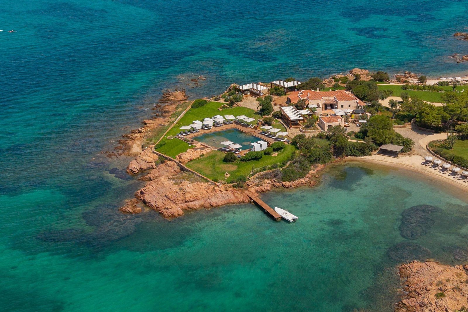 Vista aérea de resort em península com piscina cercada por espreguiçadeiras e guarda-sóis brancos. Píer de madeira com barco ancorado se estende para o mar azul-turquesa, com formações rochosas ao redor e vegetação verde no terreno.
