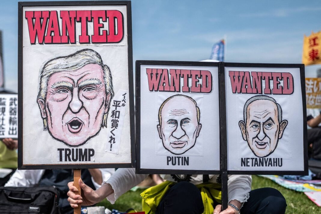 Três cartazes com caricaturas de Donald Trump, Vladimir Putin e Benjamin Netanyahu exibidos em protesto ao ar livre. Cada cartaz tem a palavra 
