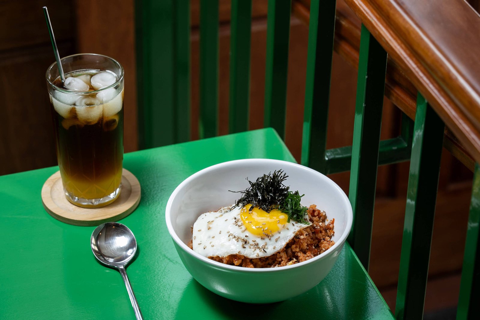 Tigela branca com arroz temperado coberto por ovo frito com gema mole e algas secas, sobre mesa verde. Ao lado, copo alto com chá gelado e gelo, com colher ao lado.
