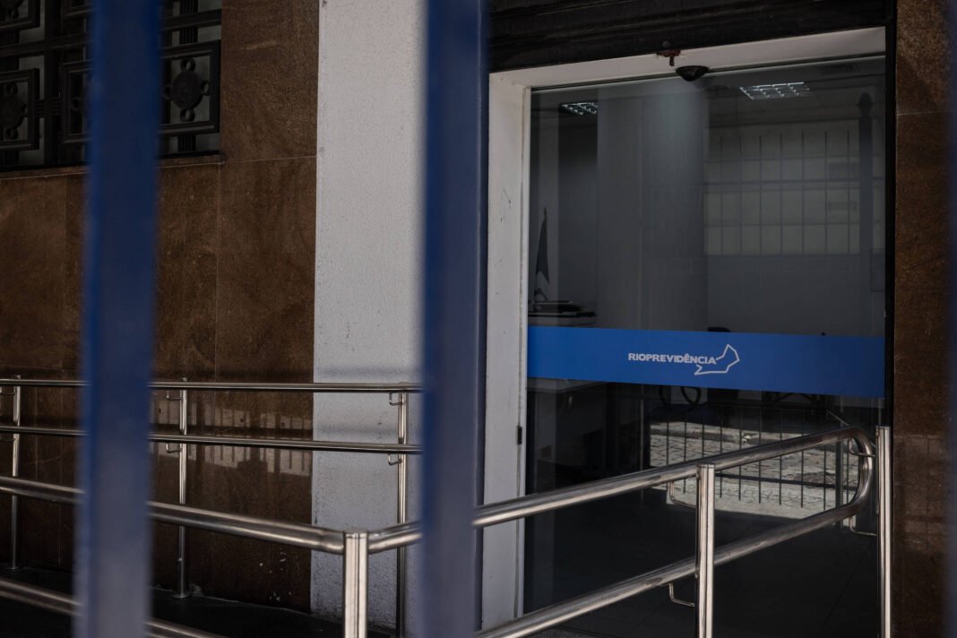 Foto mostra a entrada de prédio com rampa de acesso e corrimão de metal. A porta de vidro do prédio tem uma faixa azul com o logo do Rioprevidência.