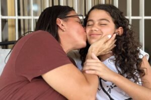 Mulher de camiseta marrom beija no rosto menina de camiseta branca com olhos fechados, sentadas em sofá com grades ao fundo.
