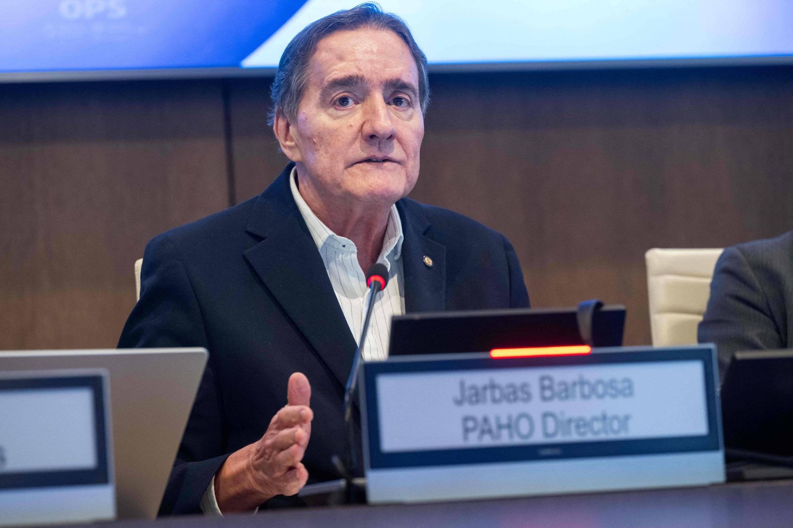 Jarbas Barbosa sentado em mesa de conferência, falando ao microfone. Placa à sua frente indica seu nome e cargo como diretor da OPAS. Fundo com painel azul e parede de madeira.